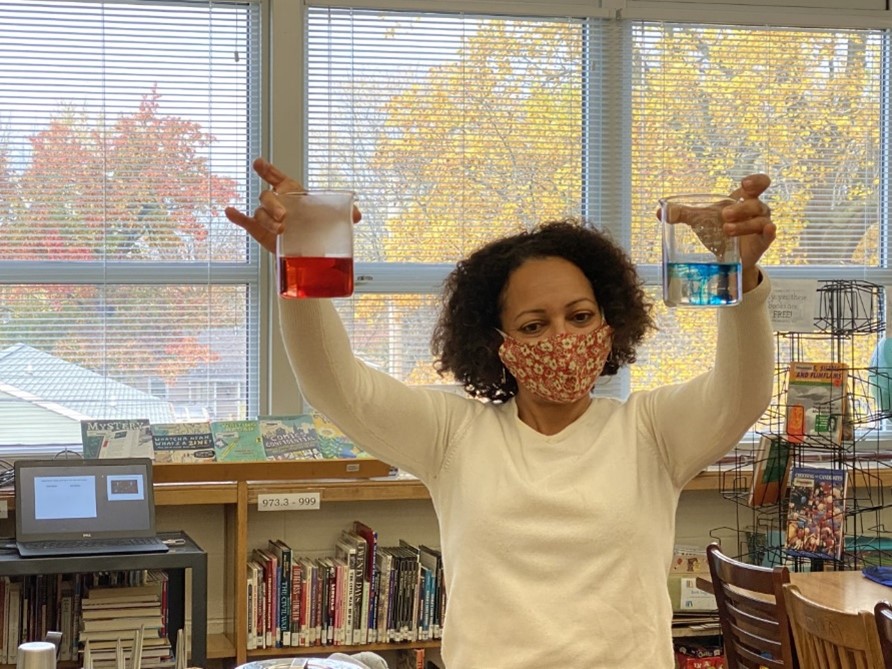 Mason holds up two beakers of water, demonstrating to students what happens when dye is dropped into steaming hot water and a frigid cold water.